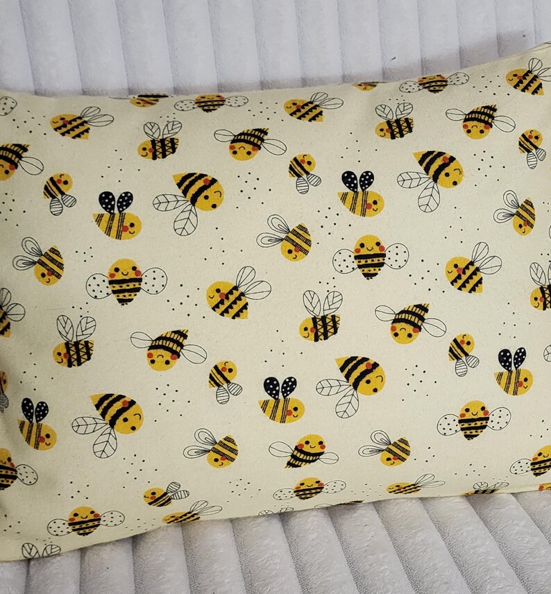 May include: Rectangular pillow with a cream-colored background covered in a pattern of yellow and black cartoon bees. The bees have smiling faces and are surrounded by small black dots.