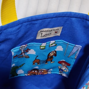 May include: A blue fabric tote bag with yellow straps featuring an interior pocket printed with cartoon characters including a dog, a cowgirl, and a potato figure. A small white label inside reads "Handmade with Love" with a red heart icon.