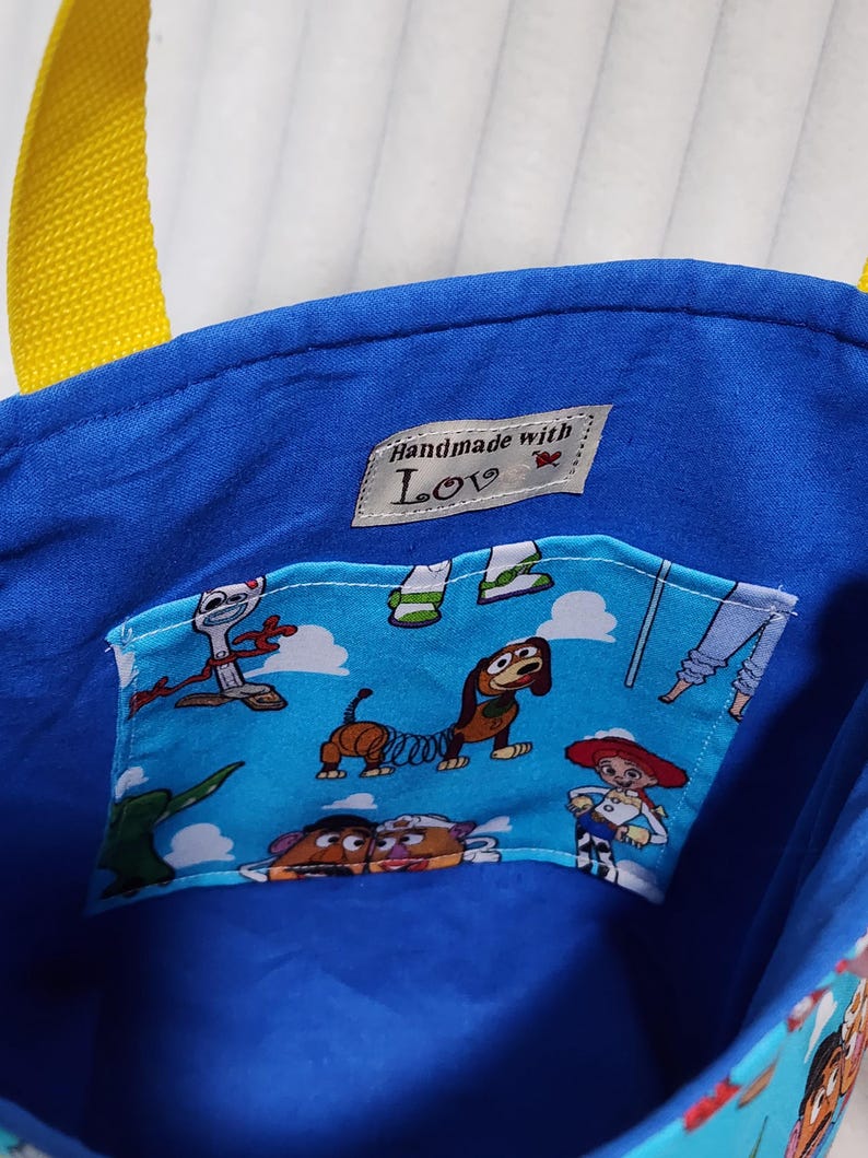 May include: A blue fabric tote bag with yellow straps featuring an interior pocket printed with cartoon characters including a dog, a cowgirl, and a potato figure. A small white label inside reads "Handmade with Love" with a red heart icon.