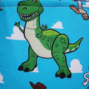 May include: A bright blue fabric featuring a repeating pattern of a green Tyrannosaurus Rex with a white belly and black claws, alongside a cowboy character wearing a brown hat, set against a background of white clouds on a blue sky.