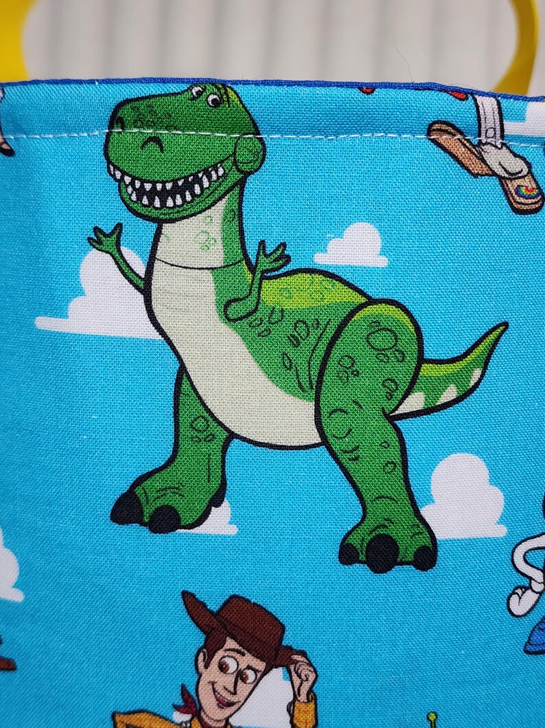 May include: A bright blue fabric featuring a repeating pattern of a green Tyrannosaurus Rex with a white belly and black claws, alongside a cowboy character wearing a brown hat, set against a background of white clouds on a blue sky.