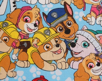 May include: A colorful fabric with a repeating pattern of cartoon dogs from the Paw Patrol television show. The dogs are wearing different colored uniforms and hats. The background is a light blue color with white paw prints.