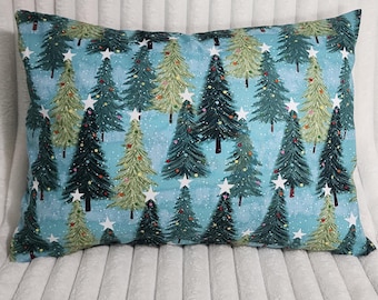 May include: A rectangular throw pillow featuring a light blue background with a repeating pattern of dark green and light green pine trees. The trees are decorated with small colorful dots and topped with white stars, set against a snowy backdrop.