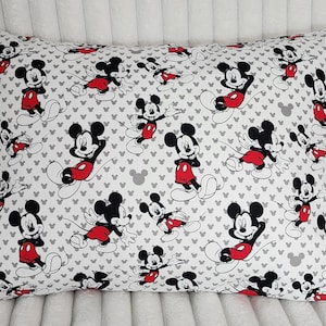 Puede incluir: Almohada rectangular con un patrón repetido de Mickey Mouse en negro, rojo y blanco sobre un fondo blanco. El diseño incluye formas de cabeza de Mickey Mouse grises. Ideal para la decoración infantil.