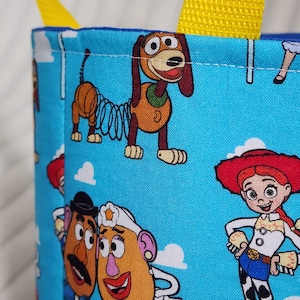 May include: A light blue fabric bag with yellow nylon straps featuring a repeating pattern of cartoon characters including a brown spring dog, a potato-shaped character with a mustache, a potato-shaped character with a white hat, and a cowgirl.
