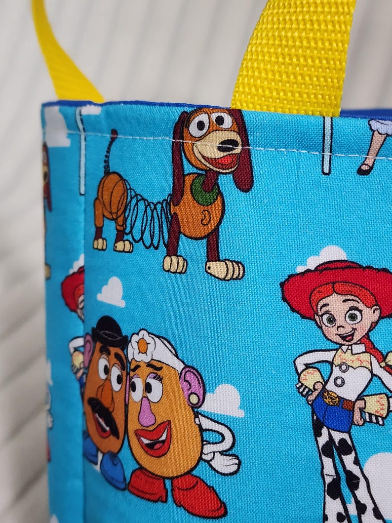 May include: A light blue fabric bag with yellow nylon straps featuring a repeating pattern of cartoon characters including a brown spring dog, a potato-shaped character with a mustache, a potato-shaped character with a white hat, and a cowgirl.