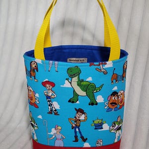 May include: A fabric tote bag featuring a light blue background with a repeating pattern of cartoon characters including a green dinosaur, a cowboy, a cowgirl, and potato figures. The bag has bright yellow handles and a solid red base.
