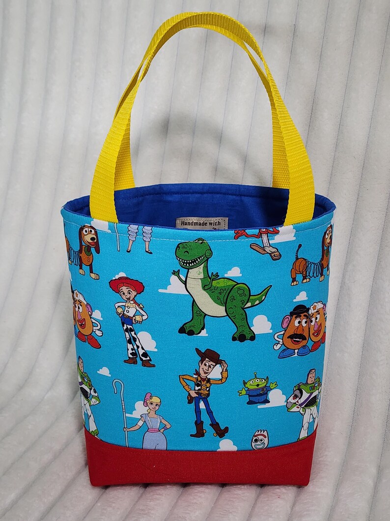 May include: A fabric tote bag featuring a light blue background with a repeating pattern of cartoon characters including a green dinosaur, a cowboy, a cowgirl, and potato figures. The bag has bright yellow handles and a solid red base.