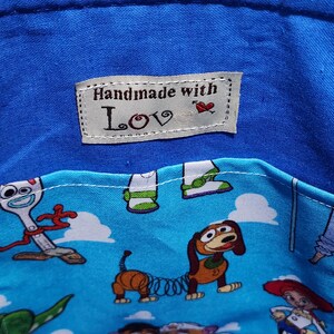 May include: A blue fabric container featuring a light blue pocket with a repeating pattern of cartoon characters including a spork, a dachshund, and a cowgirl. A white rectangular label is sewn on the interior with the text Handmade with Love.