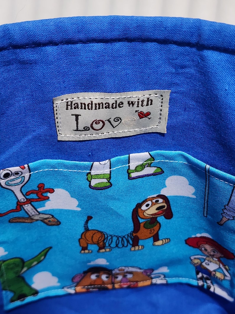 May include: A blue fabric container featuring a light blue pocket with a repeating pattern of cartoon characters including a spork, a dachshund, and a cowgirl. A white rectangular label is sewn on the interior with the text Handmade with Love.