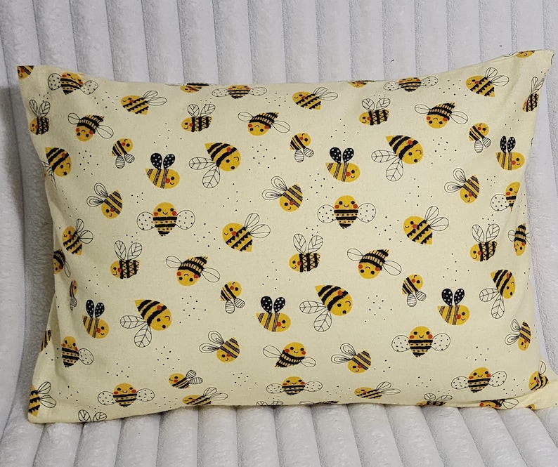 May include: Rectangular pillow with a cream-colored background and a repeating pattern of cartoon bees. The bees are yellow and black with black outlines, and have smiling faces. The pillow is a decorative item.