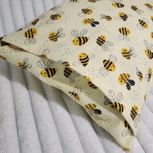 May include: A square pillow with a cream-colored background and a repeating pattern of cartoon bees. The bees are yellow and black with smiling faces and black and white wings. The pillow is made of a soft fabric.