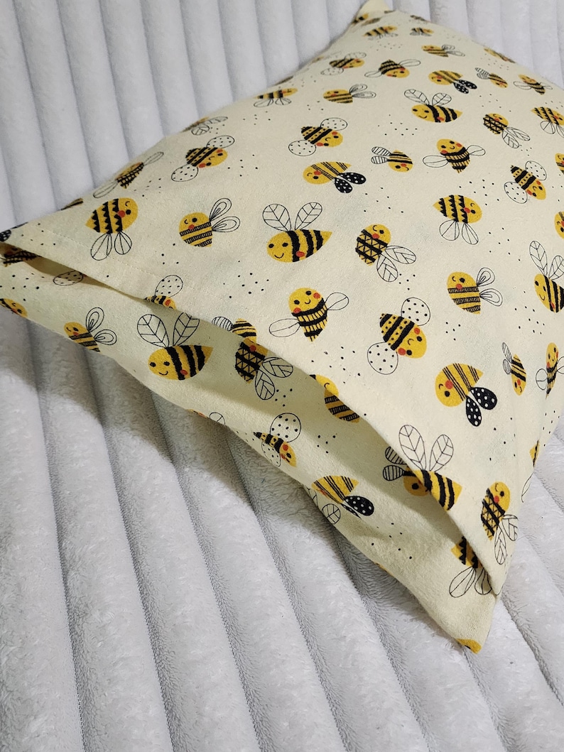 May include: A square pillow with a cream-colored background and a repeating pattern of cartoon bees. The bees are yellow and black with smiling faces and black and white wings. The pillow is made of a soft fabric.