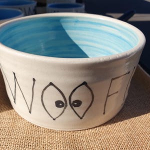 May include: White ceramic pet bowl with a light blue interior. The word "WOOF" is written in black, with two stylized eye shapes. The bowl is likely for a dog or other pet.