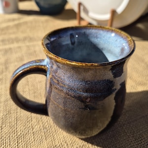 May include: A ceramic mug with a rounded body and a curved handle. The exterior features a mottled glaze in shades of deep brown, charcoal, and hints of blue. The interior of the mug has a smooth, light blue glaze, while the rim is finished in a warm, earthy brown tone.