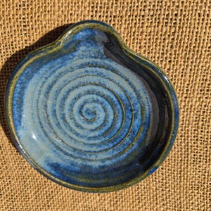 May include: A round ceramic spoon rest featuring a glossy glaze in shades of deep blue and light blue. The surface displays a prominent spiral pattern etched into the centre, with a dark green rim accentuating the irregular, organic edge of the piece.