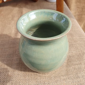 May include: A light green ceramic vase with a wide opening and rounded body. The vase has a smooth, glazed finish and a slightly textured surface. The vase is suitable for holding flowers or as a decorative item.