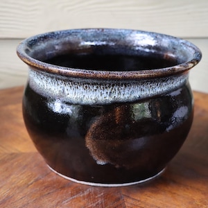 May include: A handmade ceramic pot with a glossy, dark brown exterior. The rim has a band of blue and white glaze, and the interior is dark. The pot sits on a wooden surface.