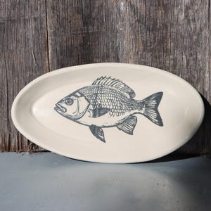 May include: An oval-shaped ceramic dish with a smooth off-white finish. The centre of the dish features a detailed, dark grey graphic illustration of a fish with visible scales, fins, and eye detail, centred on the surface of the plate.