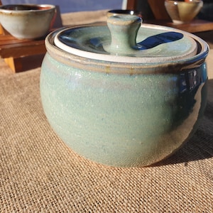 May include: A round, ceramic lidded jar with a glossy, light sage green glaze. The jar features a smooth, bulbous body and a matching lid with a small, centred knob handle. The glaze shows subtle variations in tone, with hints of blue and brown.