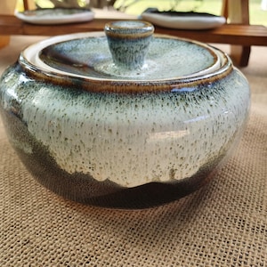 May include: A ceramic lidded jar with a speckled glaze in shades of blue, white, and brown. The jar has a rounded shape with a matching lid and a small knob on top. The jar is sitting on a hessian surface.