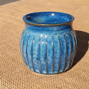 May include: A handmade ceramic vase with a glossy, textured finish. The vase is a vibrant blue with vertical grooves and a contrasting rim. The round shape and unique design make it a decorative piece for any home.