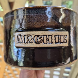 May include: A cylindrical ceramic bowl with a glossy, dark brown glaze. A rectangular, unglazed beige plaque is centred on the front, featuring the name ARCHIE embossed in bold, capital letters with a dark outline around each character.