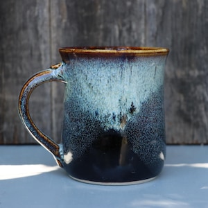 May include: A ceramic mug featuring a reactive glaze with a deep navy blue base that transitions into a lighter, speckled mint green toward the rim. The mug has a curved handle and a smooth, glossy finish with a dark brown rim detail.