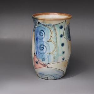 May include: A hand-painted ceramic vase with a cream base and colourful abstract designs. The vase features blue, teal, and peach shapes with black outlines and spiral details. The rim is a warm brown colour.