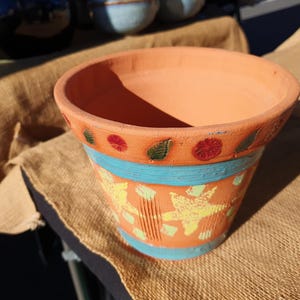 May include: A terracotta flower pot featuring a wide rim decorated with small green leaf and red flower motifs. The body of the pot is painted with a horizontal blue band and yellow starfish designs against an orange background with vertical texture.