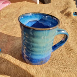 May include: A handmade ceramic mug with a blue and teal glaze. The mug has a rounded shape with a handle and a brown rim. The mug is sitting on a burlap surface.