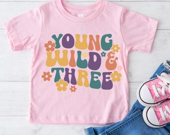 May include: Pink short-sleeved t-shirt with a retro-style graphic that says "Young Wild & Three" in colorful letters with flowers around the text.