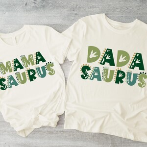 May include: Two white t-shirts with green and brown dinosaur-themed text. The shirt on the left says "Mama Saurus" and the shirt on the right says "Dada Saurus".