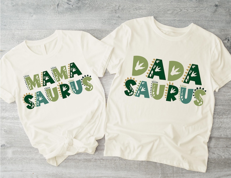 May include: Two white t-shirts with green and brown dinosaur-themed text. The shirt on the left says "Mama Saurus" and the shirt on the right says "Dada Saurus".