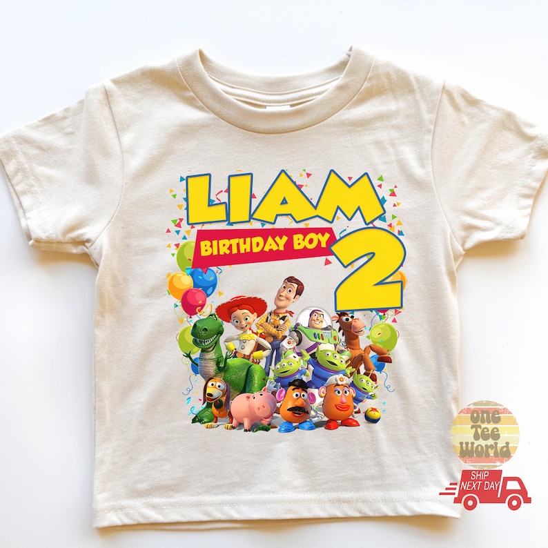 May include: Cream-colored t-shirt featuring the name "Liam" in yellow block letters with a blue outline, and the number "2". The shirt also displays the text "Birthday Boy" and a colorful graphic of Toy Story characters. Balloons and confetti are also present.