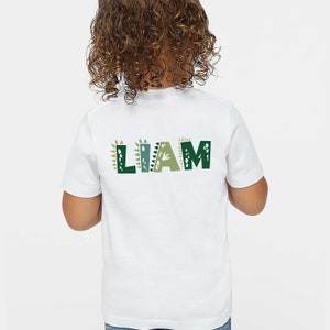 May include: White t-shirt with the name "LIAM" printed on the back in green, brown, and white letters with dinosaur-themed designs.