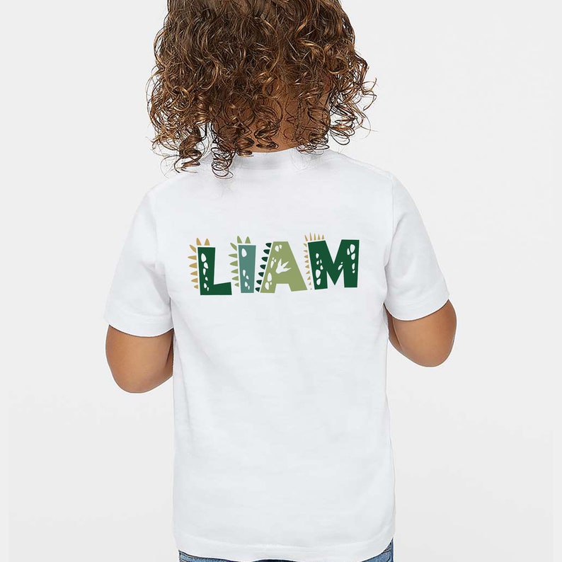 May include: White t-shirt with the name "LIAM" printed on the back in green, brown, and white letters with dinosaur-themed designs.