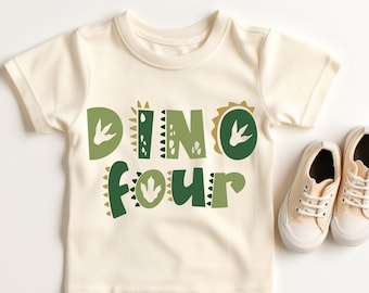 Op de afbeelding: Een crèmekleurig T-shirt met korte mouwen met de woorden "Dino Four" in groene en gouden letters. De letters zijn versierd met dinosaurusvoetafdrukken en andere patronen.
