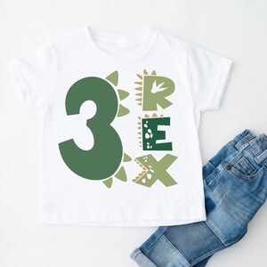 May include: White t-shirt with a green number 3 and the word "REX" in green letters. The letters and number are surrounded by brown dinosaur scales.