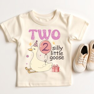 May include: Cream-colored toddler t-shirt with a whimsical goose graphic. The design includes the text "TWO silly little goose" in purple, a goose wearing a party hat, and a pink balloon. A pair of beige sneakers are next to the shirt.