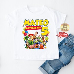 May include: White t-shirt featuring a colorful graphic of Toy Story characters with the name "Mateo" and the number "5" in yellow. The text "BIRTHDAY BOY" is also included. The shirt is paired with blue jeans.