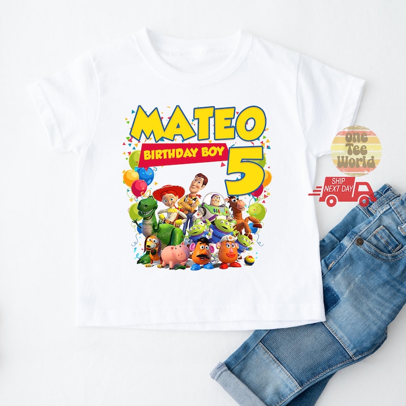 May include: White t-shirt featuring a colorful graphic of Toy Story characters with the name "Mateo" and the number "5" in yellow. The text "BIRTHDAY BOY" is also included. The shirt is paired with blue jeans.