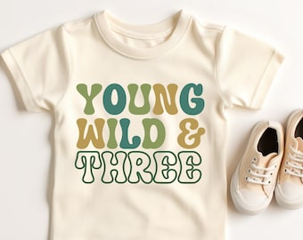 May include: A cream colored t-shirt with the text "YOUNG WILD & THREE" in green and gold retro font.