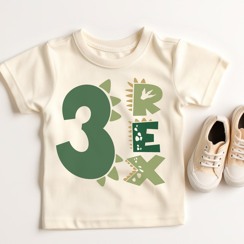 May include: A cream colored t-shirt with a green number "3" and the word "REX" in green and brown letters. The letters are decorated with dinosaur-like spikes and footprints.