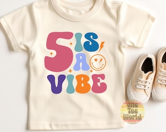 Peut inclure: T-shirt crème avec l'inscription "Sis is a Vibe" dans une police rétro colorée. Le motif comprend un smiley et un éclair. Une paire de baskets beiges est à côté du t-shirt.