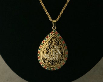 May include: Gold necklace with a teardrop-shaped pendant featuring an intricate design and the word "Allah" in Arabic script. The pendant is adorned with clear and colored rhinestones.