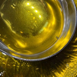 May include: Close-up of a stainless steel bowl filled with golden-yellow liquid, possibly oil. The liquid reflects light, creating bright spots and a shimmering effect. Small bubbles are visible near the edges of the bowl, adding texture to the image.