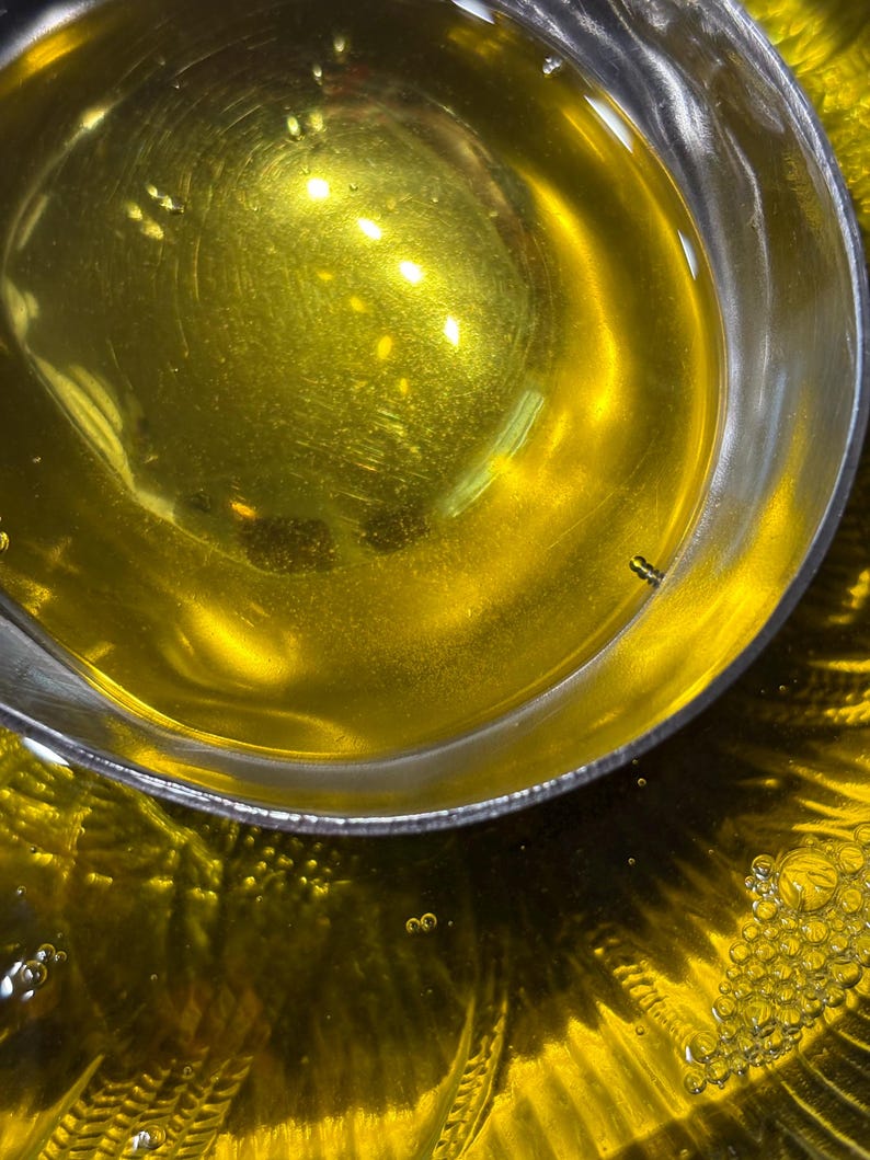 May include: Close-up of a stainless steel bowl filled with golden-yellow liquid, possibly oil. The liquid reflects light, creating bright spots and a shimmering effect. Small bubbles are visible near the edges of the bowl, adding texture to the image.