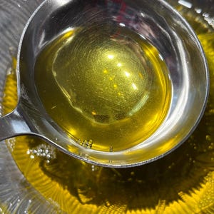 May include: A stainless steel ladle filled with golden liquid, held over a glass bowl containing the same liquid. The liquid's surface reflects light, with tiny bubbles present. The image is a close-up, emphasizing the texture and colour.