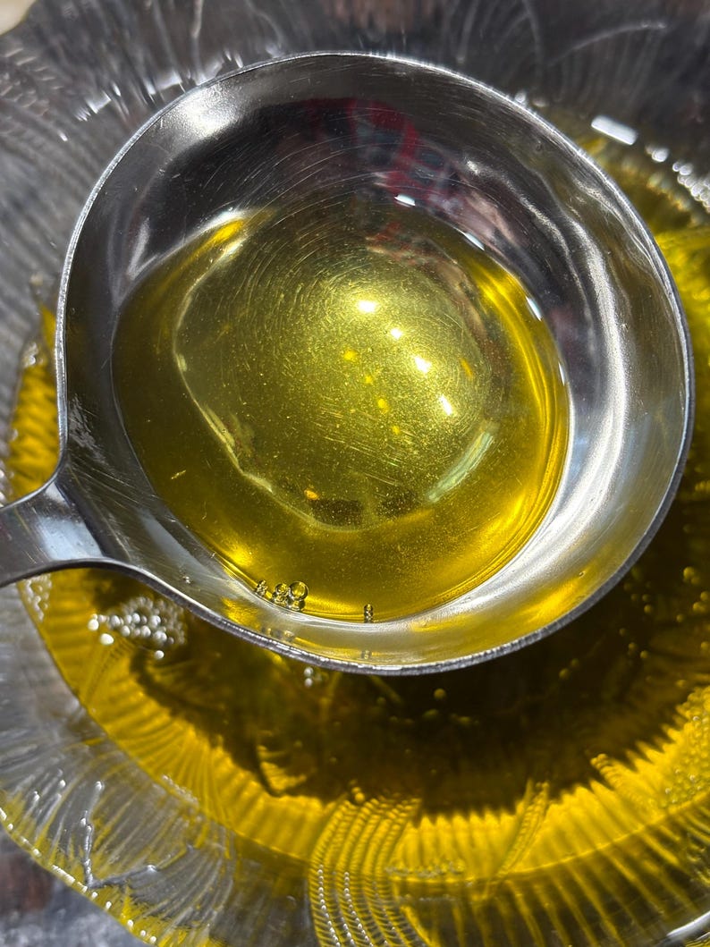 May include: A stainless steel ladle filled with golden liquid, held over a glass bowl containing the same liquid. The liquid's surface reflects light, with tiny bubbles present. The image is a close-up, emphasizing the texture and colour.